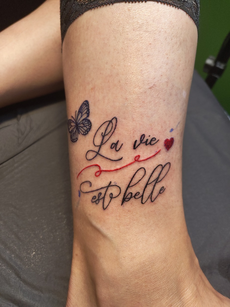 Skript-Tattoo mit dem Text 'La vie est belle' auf dem Knöchel mit einem Schmetterling und einem roten HErzen